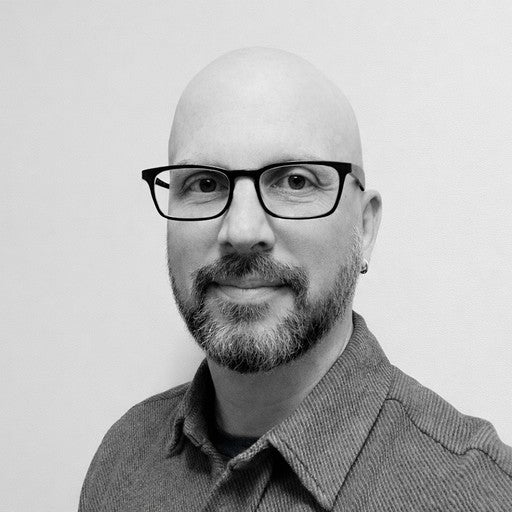 Bald man with glasses and a beard, wearing a collared shirt, smiling at the camera in black and white.