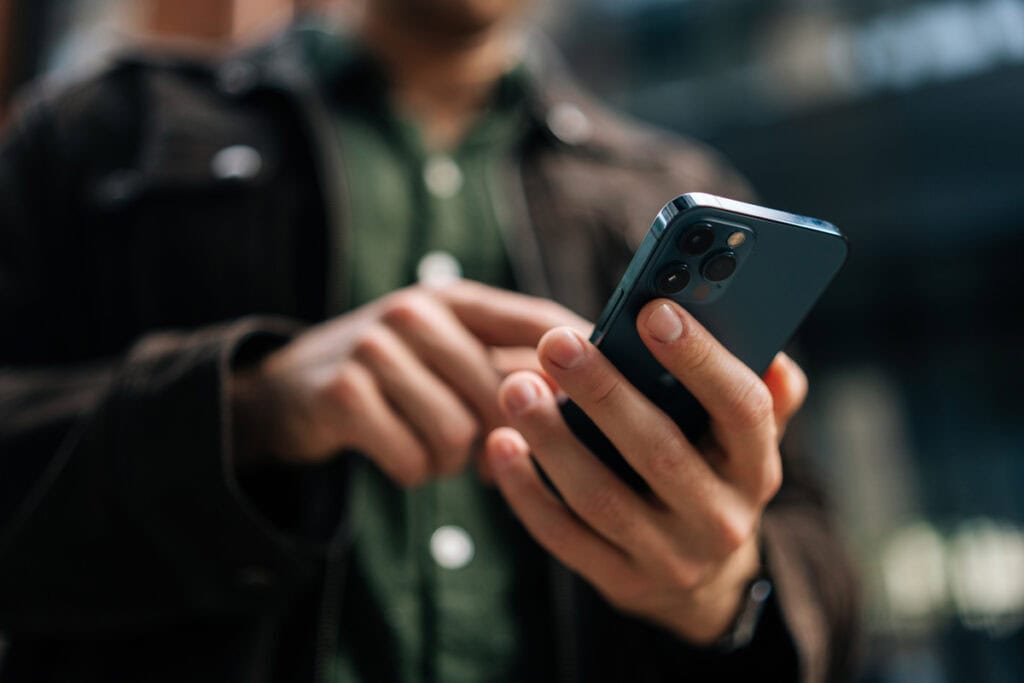 A person using a smartphone outdoors, wearing a green shirt and dark jacket, blurred background.