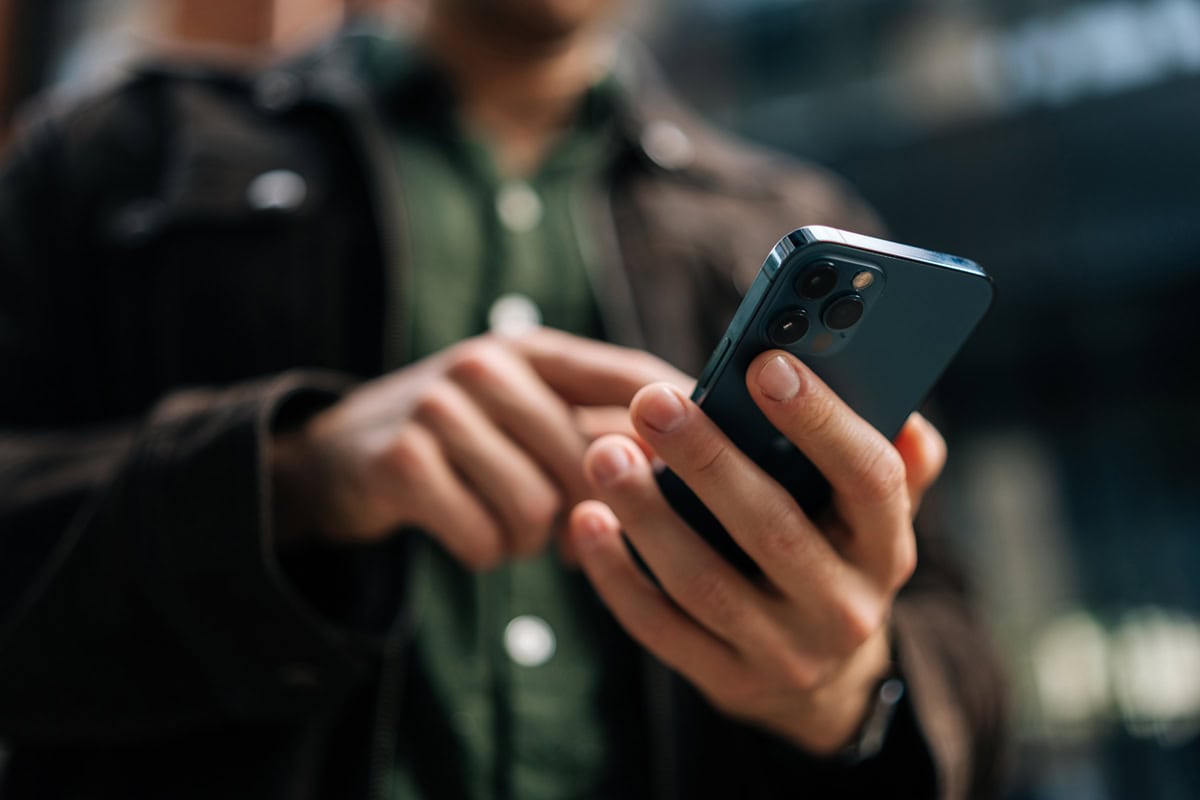 A person using a smartphone outdoors, wearing a green shirt and dark jacket, blurred background.