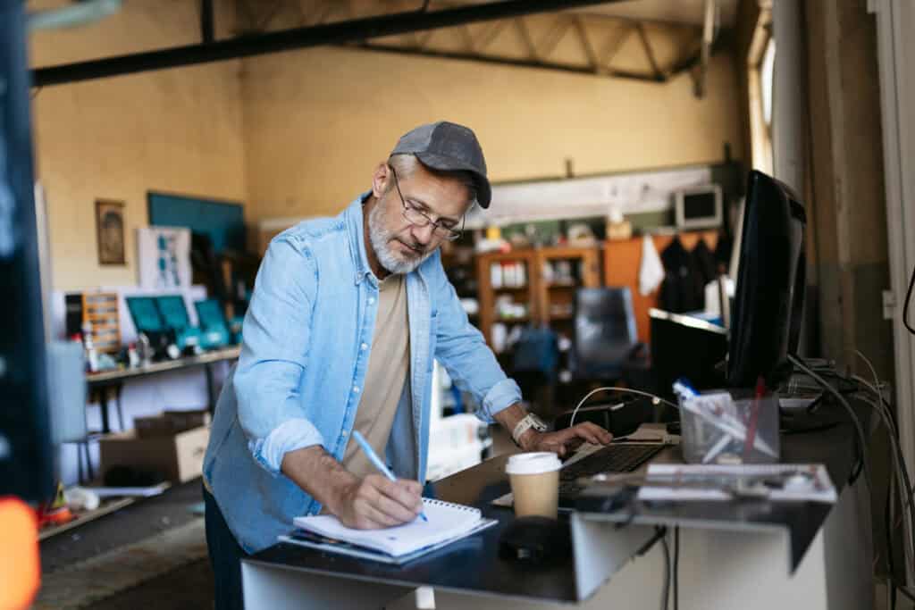 Older man in a cap writes in a notebook while working at a desk with a computer in a workshop.