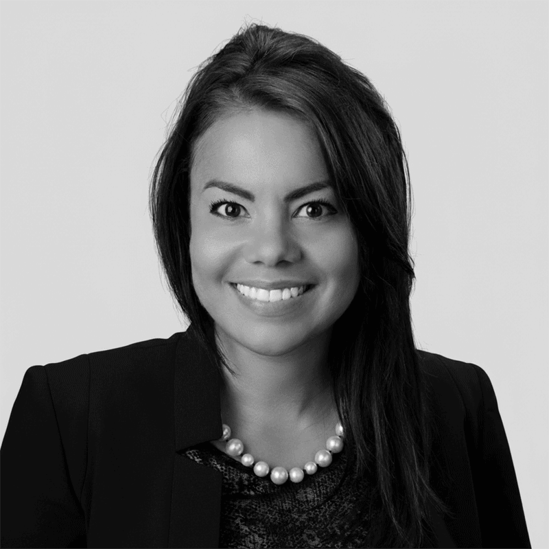 Smiling woman with long hair, wearing a blazer, pearl necklace, and dark top, on a plain background.