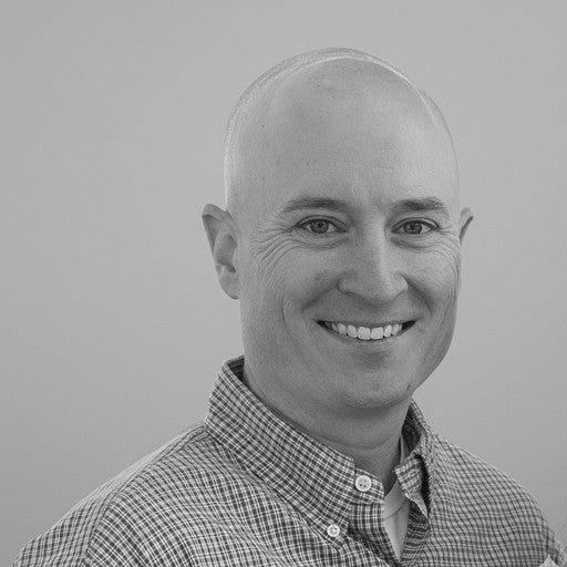 Bald man in a checked shirt smiling at the camera against a plain background.