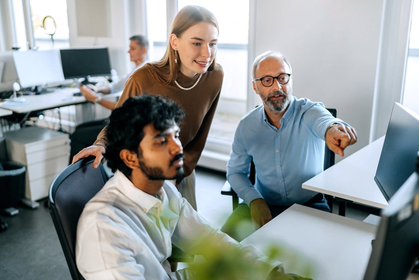Three colleagues collaborate at a desk in a bright office, with one person pointing at a computer screen while the others watch attentively.