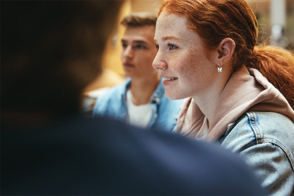 A young woman with red hair and freckles smiles, sitting next to a young man, both appearing to listen attentively in a casual indoor setting.